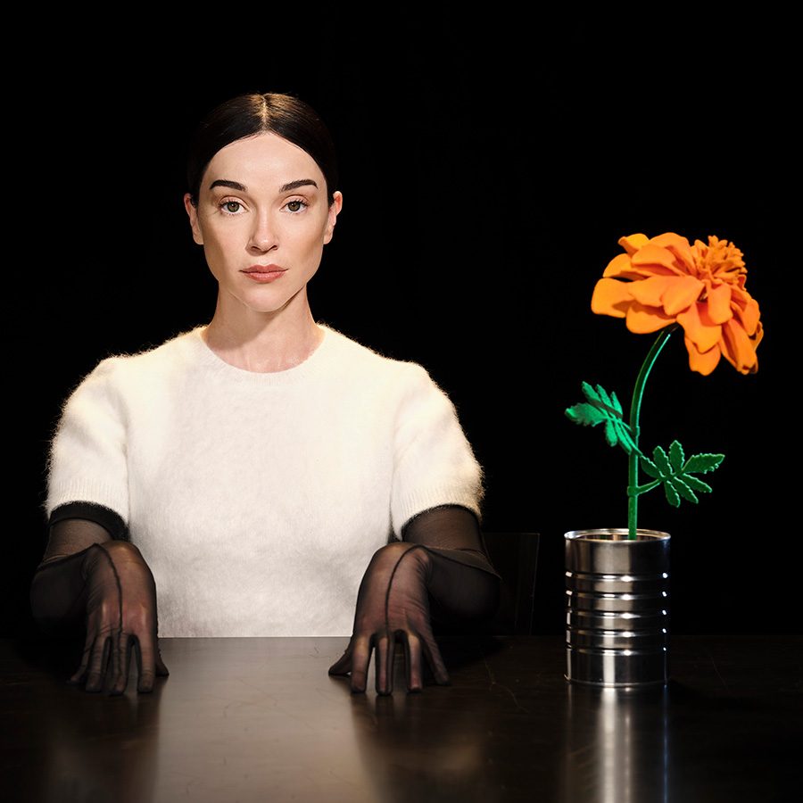 American performer St. Vincent pictured seated with an orange flower against a black background.